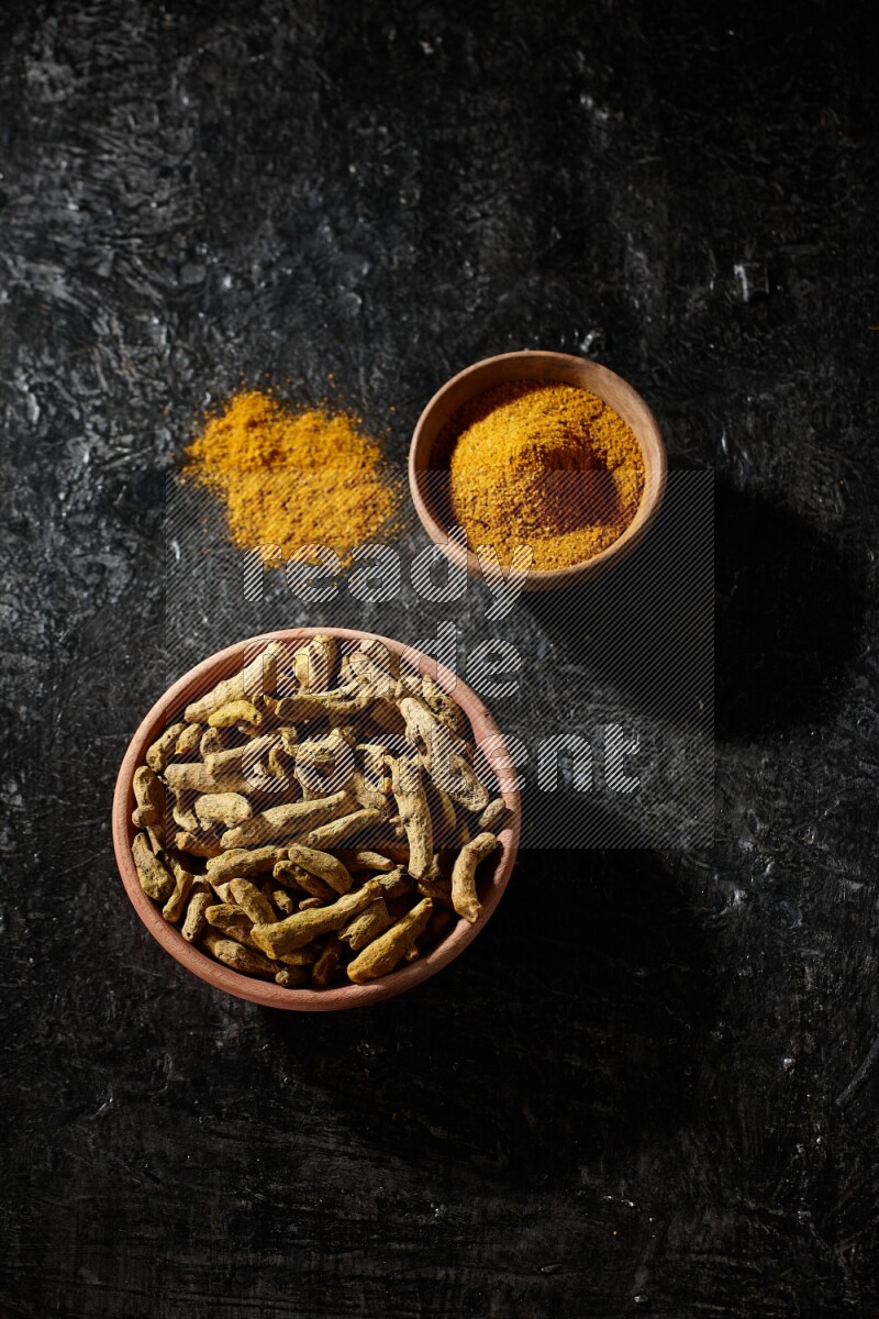 2 wooden bowls, one full of turmeric powder and the other full of dried turmeric whole fingers on a textured black flooring