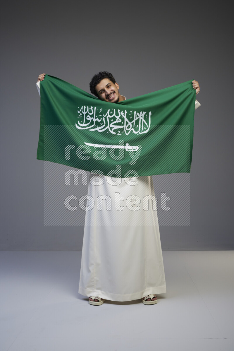 A Saudi man standing wearing thob holding big Saudi flag on gray background