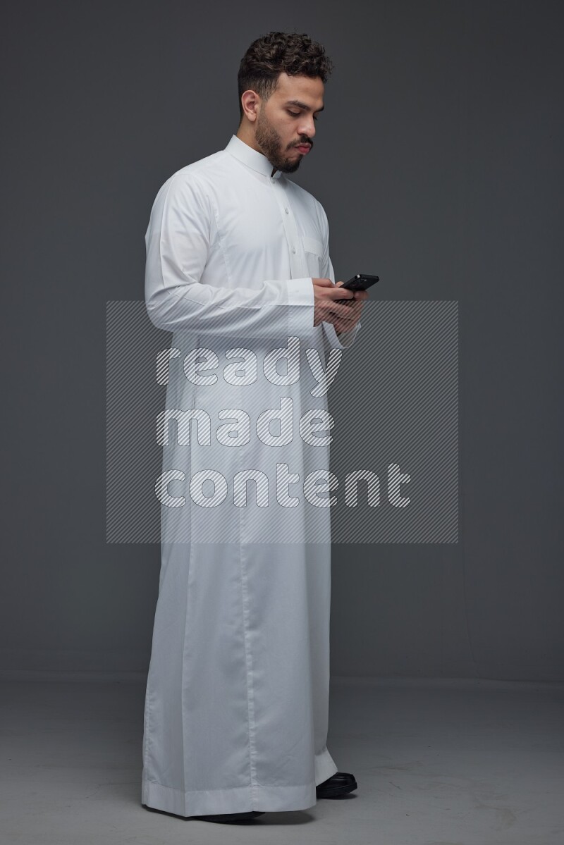 A Saudi man wearing Thobe standing and using his phone eye level on a gray background