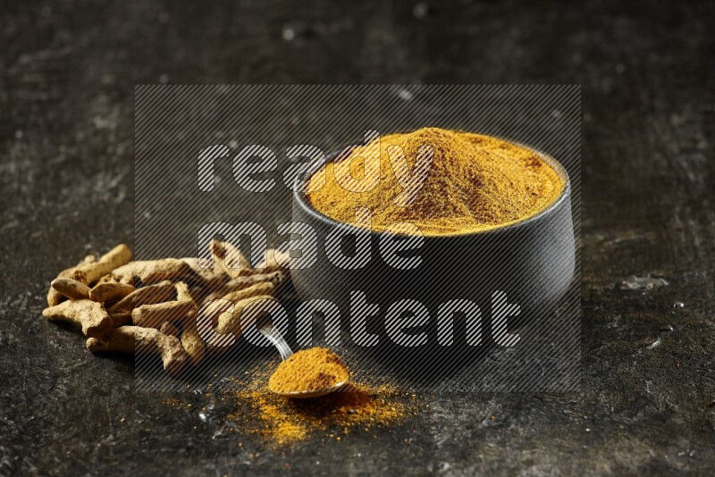 A pottery black bowl and a metal spoon full of turmeric powder with dried turmeric fingers on a textured black flooring