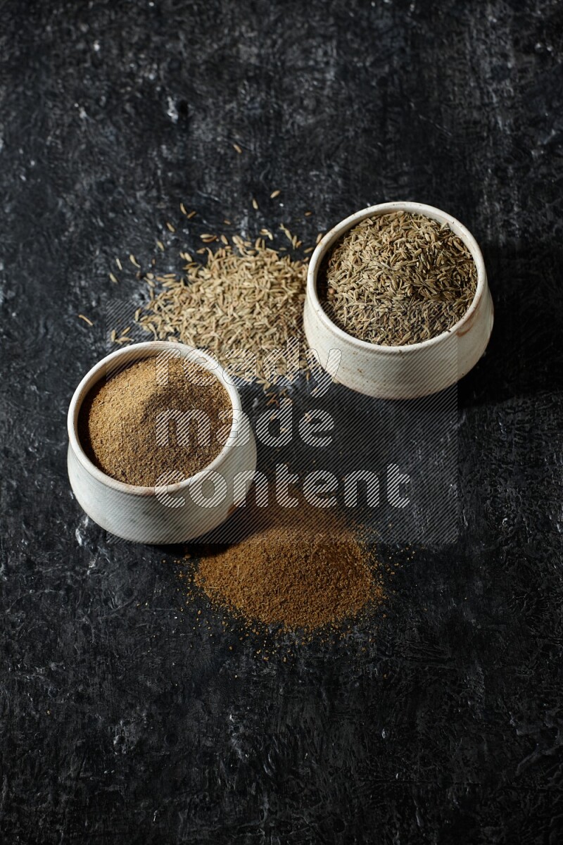 وعاءين بيج، أحدهما ممتلئ ببودرة الكمون والآخر ممتلئ بحبوب الكمون، على خلفية سوداء