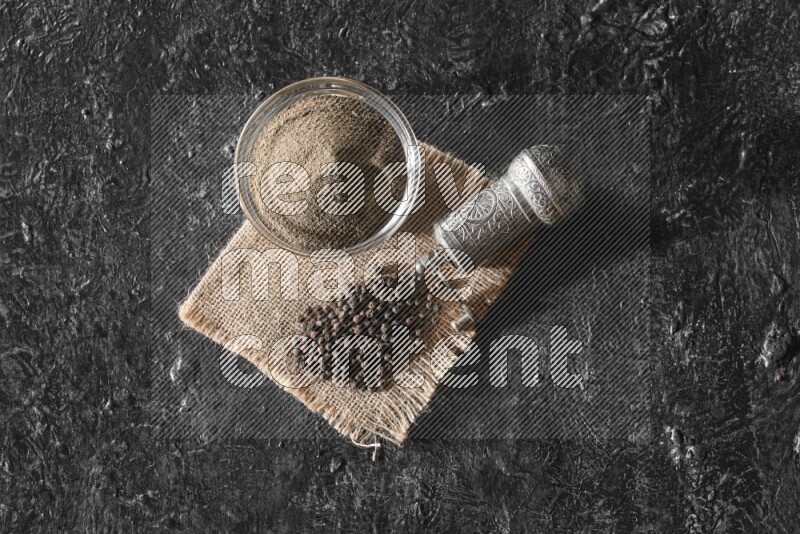 A glass bowl full of black pepper powder and black pepper beads on burlap fabric with a turkish metal pepper grinder on textured black flooring