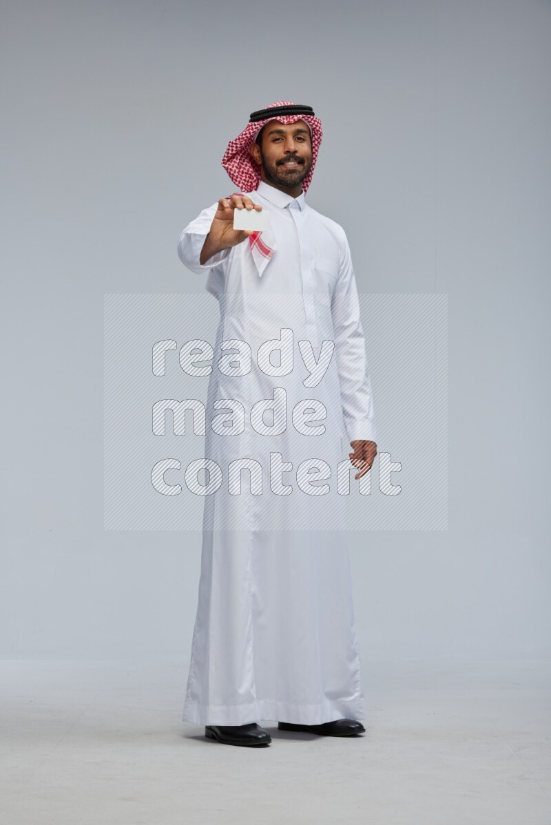 Saudi man wearing thob and shomag standing holding ATM card on gray background