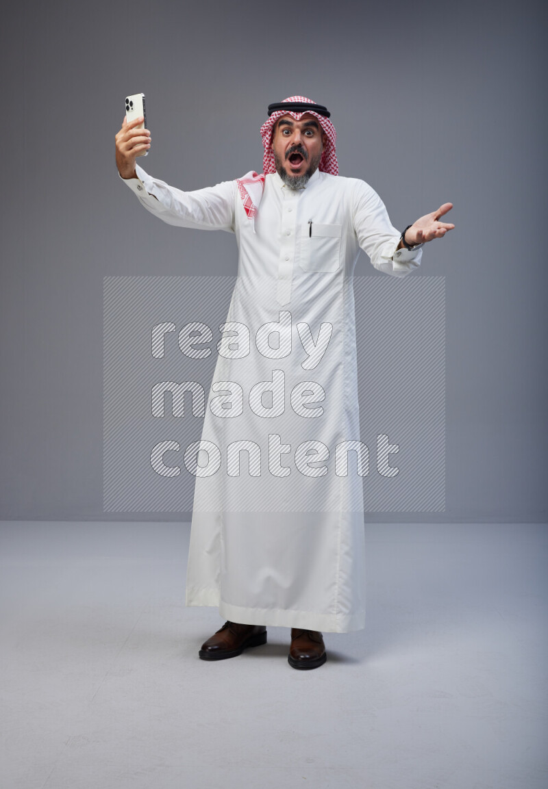 Saudi man Wearing Thob and red Shomag standing taking selfie on Gray background
