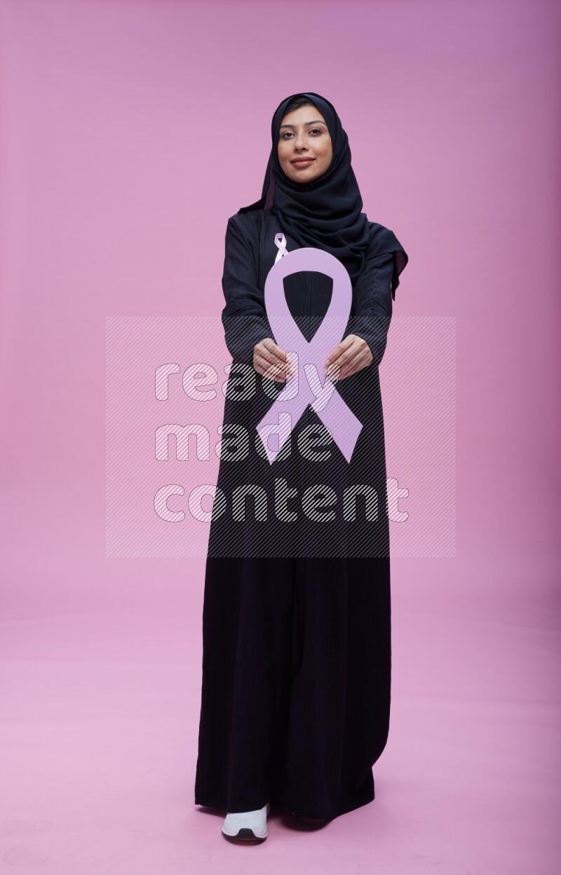 Saudi woman wearing pink ribbon on abaya standing holding awareness ribbon on plain pink background
