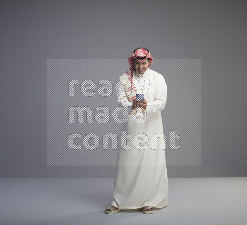 A  Saudi man standing wearing thob and red shomag texting on phone on gray background
