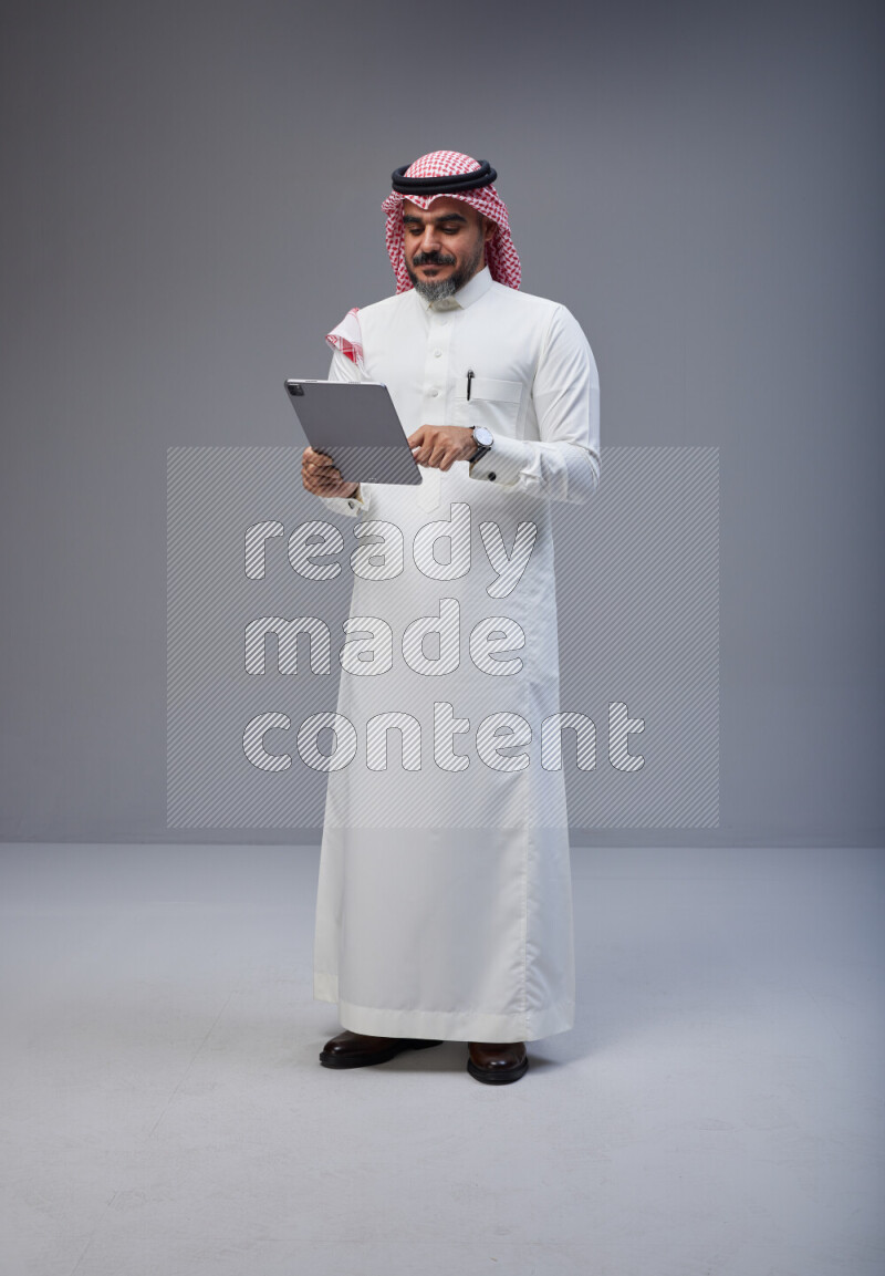 Saudi man Wearing Thob and red Shomag standing working on tablet on Gray background
