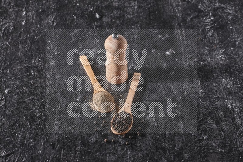 Black pepper powder and black pepper beads in wooden spoons and wooden grinder on a textured black flooring