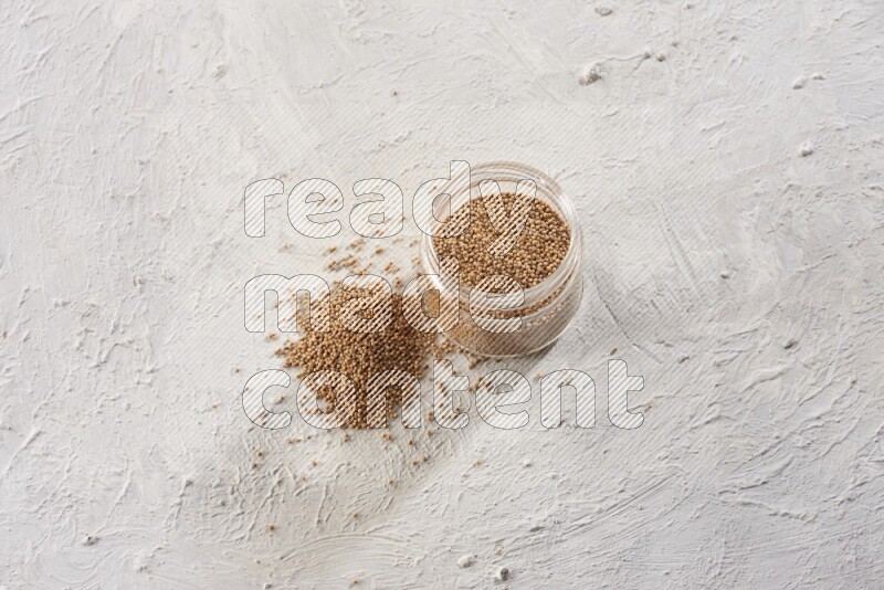A glass jar full of mustard seeds and more seeds spread on a textured white flooring