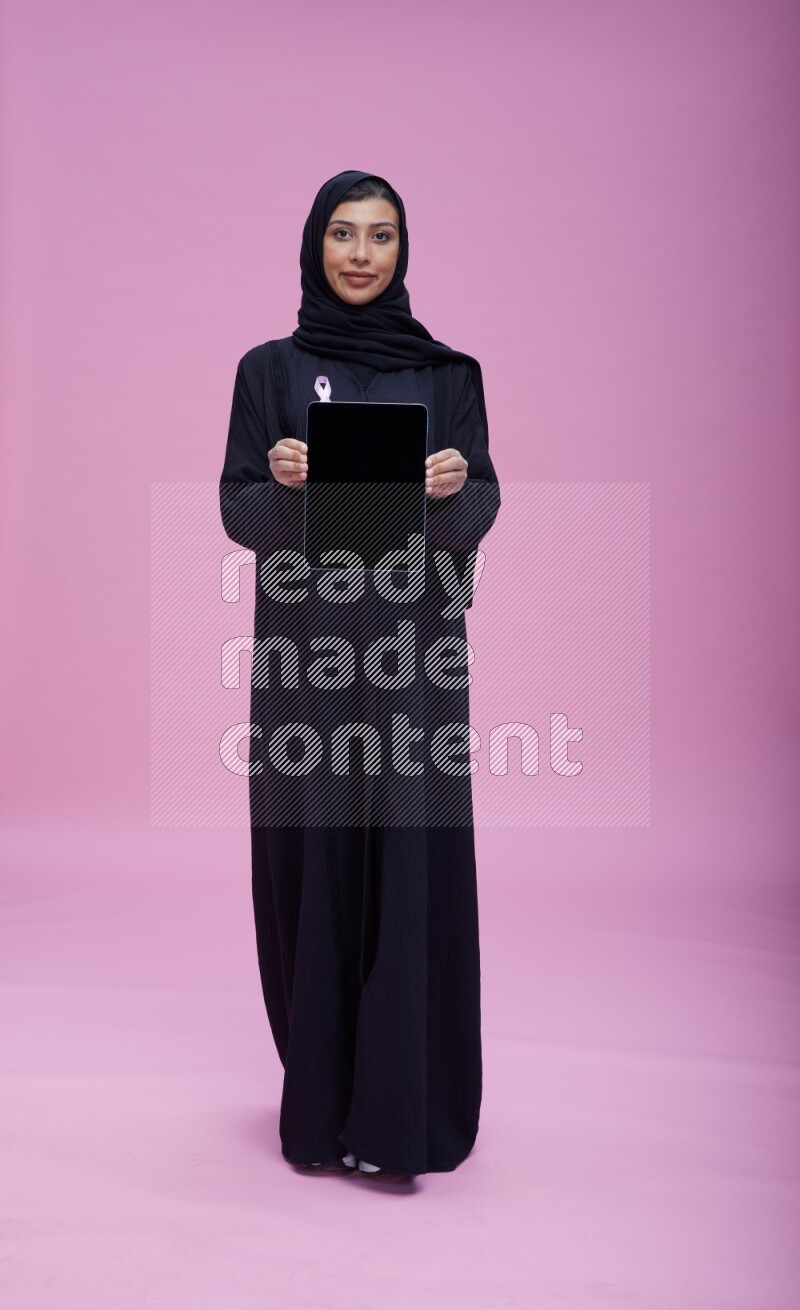 Saudi woman wearing pink ribbon on Abaya standing showing a tablet to camera on pink background