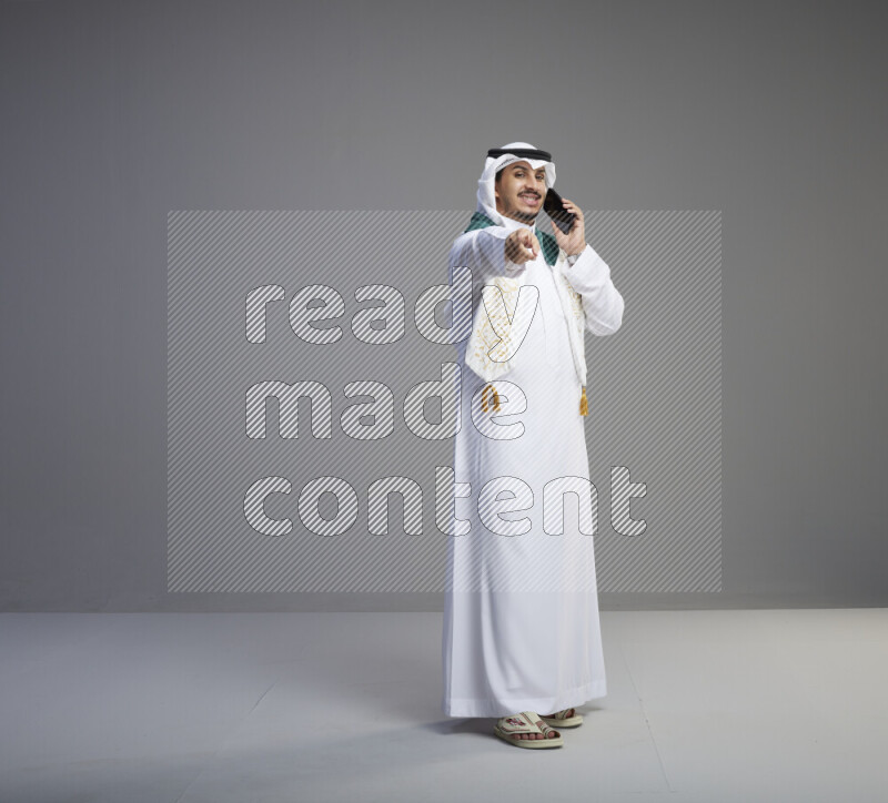 A saudi man standing wearing thob and white shomag with flag scarf on his neck talking on phone on gray background