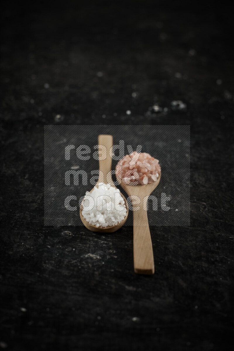 2 wooden spoons filled with fine and coarse salt on black background