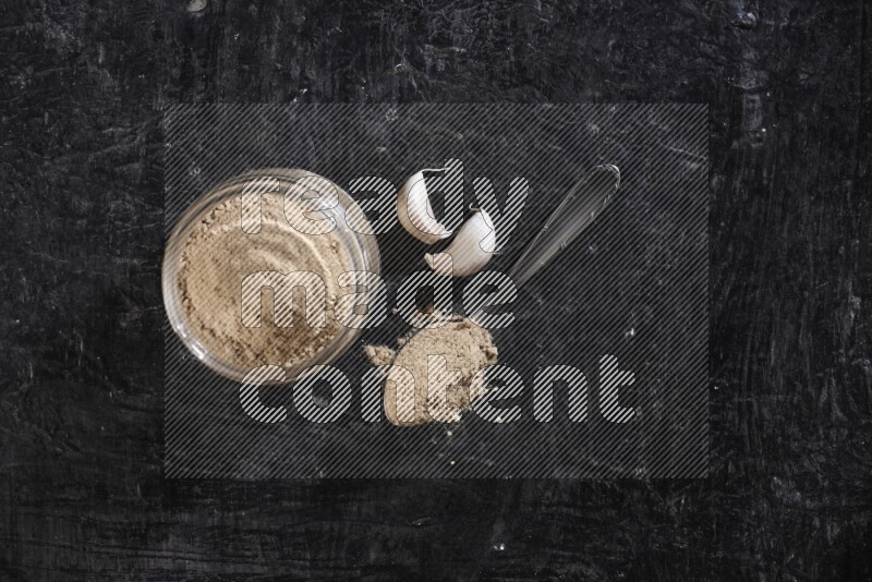 A glass jar full of garlic powder with a metal spoon full of the powder on a textured black flooring