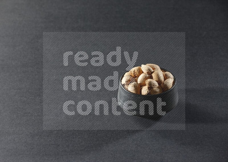 A black pottery bowl full of almonds on a black background in different angles