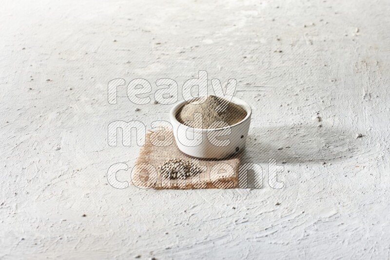 White pottery bowl full of white pepper powder set on a burlap piece of fabric with pepper beads and wooden pepper grinder on textured white flooring