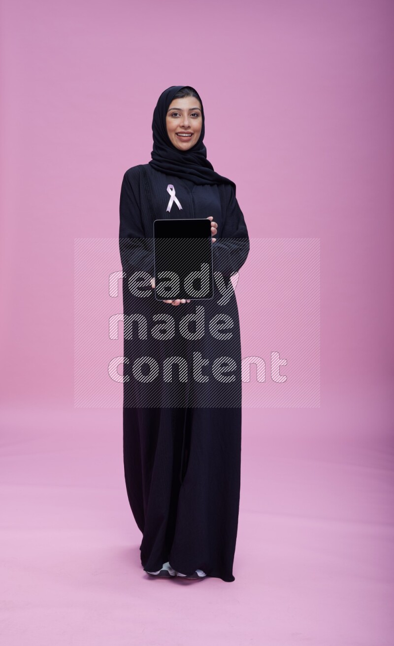 Saudi woman wearing pink ribbon on Abaya standing showing a tablet to camera on pink background