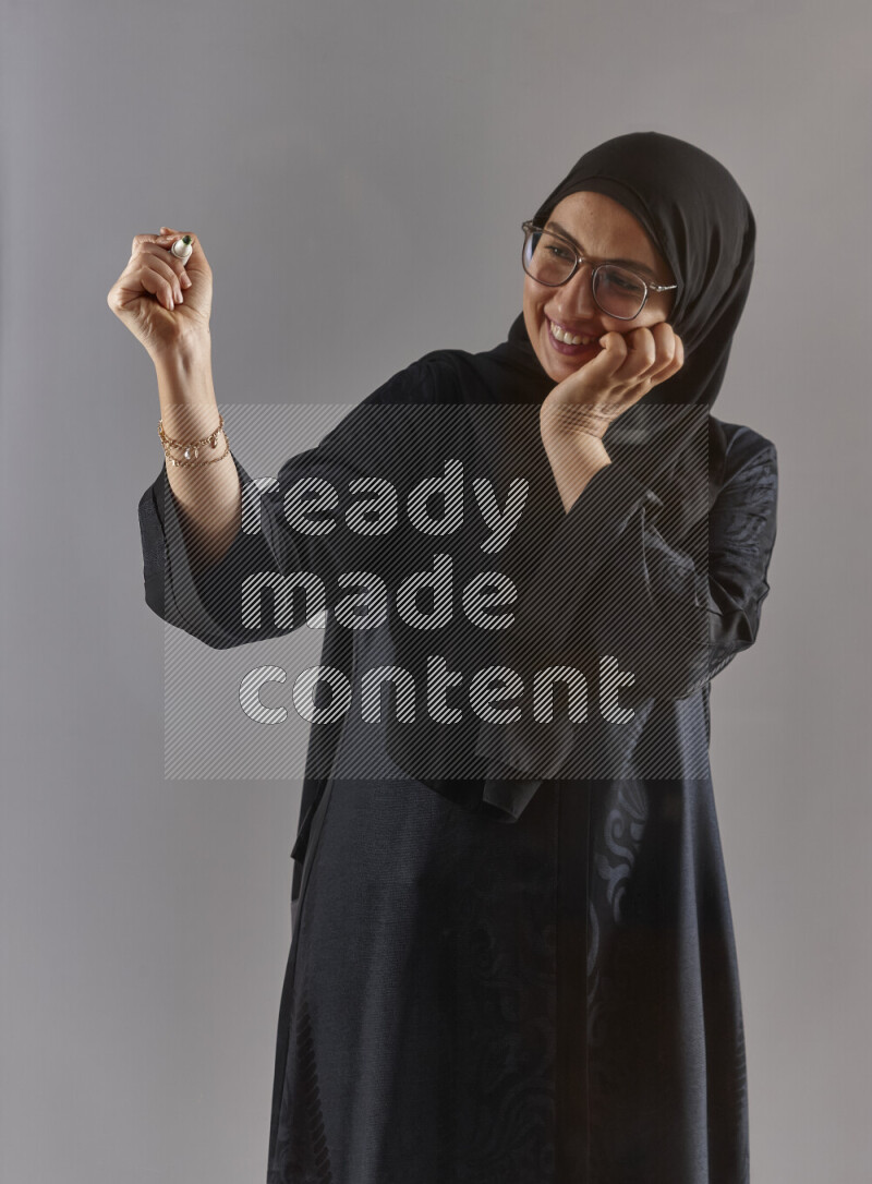 Woman in black abaya, hijab and glasses holding a marker pen to write on a transparent board with different reactions and expressions on grey background