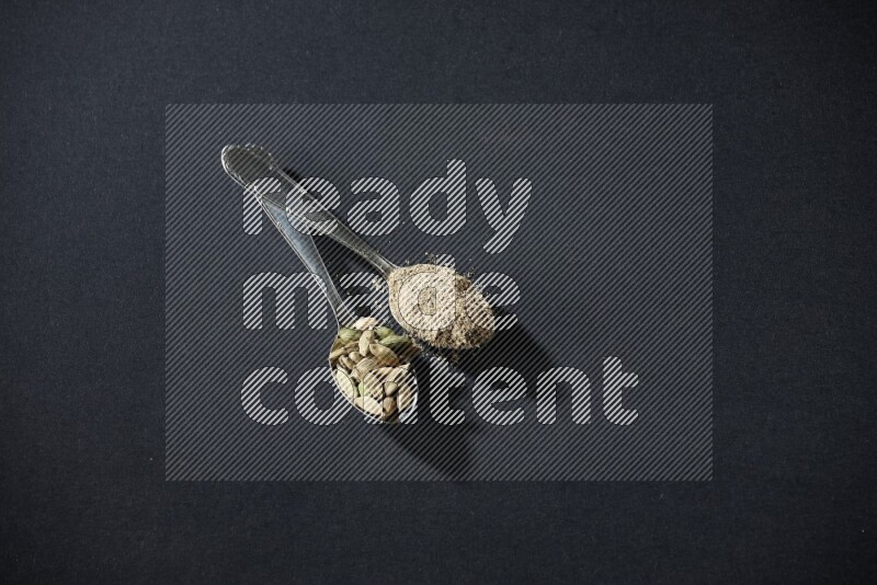 2 Metal spoons full of cardamom powder and cardamom seeds on black flooring