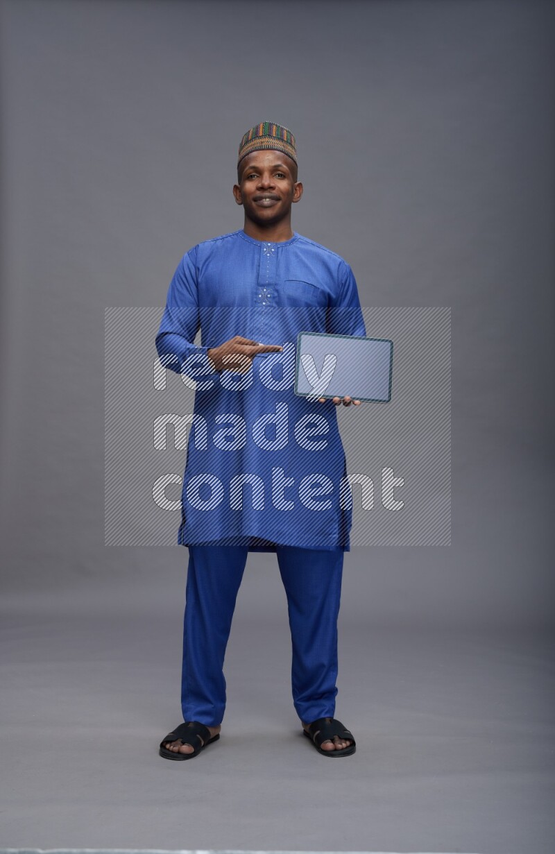 Man wearing Nigerian outfit standing showing tablet to camera on gray background
