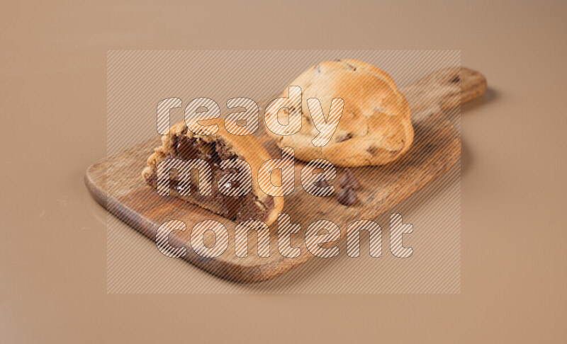 a chocolate chip cookie with another one cut in half on a wooden cutting board on a brown background