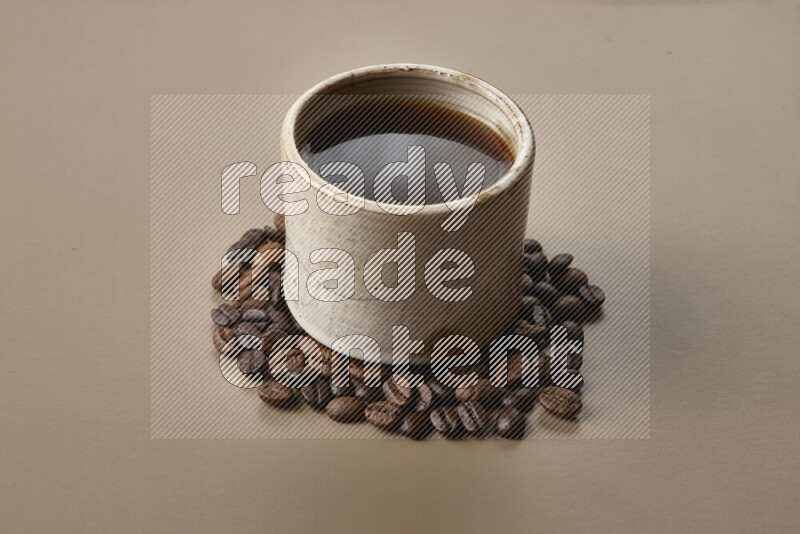 A beige pottery cup of coffee surrounded by roasted coffee beans on beige background