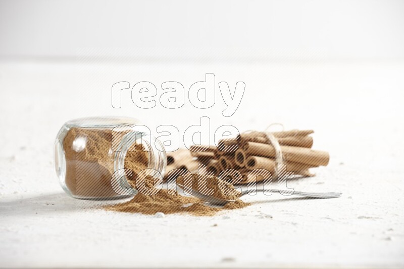 Flipped herbs glass jar full of cinnamon powder with a metal spoon full of powder and cinnamon sticks on a textured white background