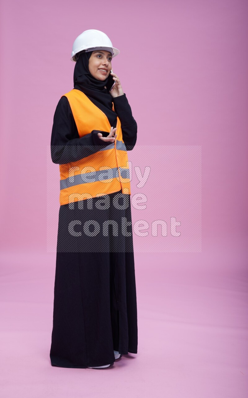 Saudi woman wearing Abaya with engineer vest and helmet standing talking on phone on pink background