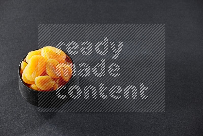 A black pottery bowl full of dried apricots on a black background in different angles