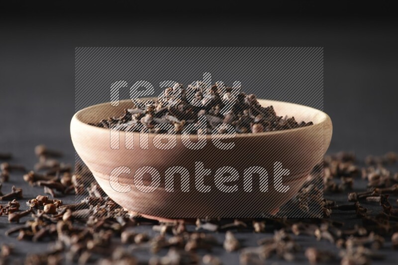 A wooden bowl full of cloves with spreaded whole cloves on a black flooring