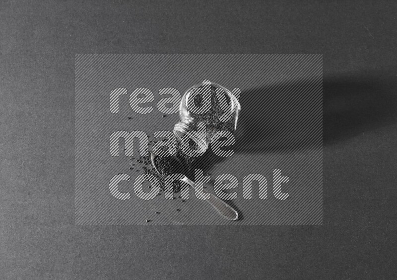 وعاء بهارات زجاجي مقلوب ممتلئ ببذور حبة البركة والبذور تناثرت منه مع ملعقة معدنية ممتلئة على أرضية سوداء