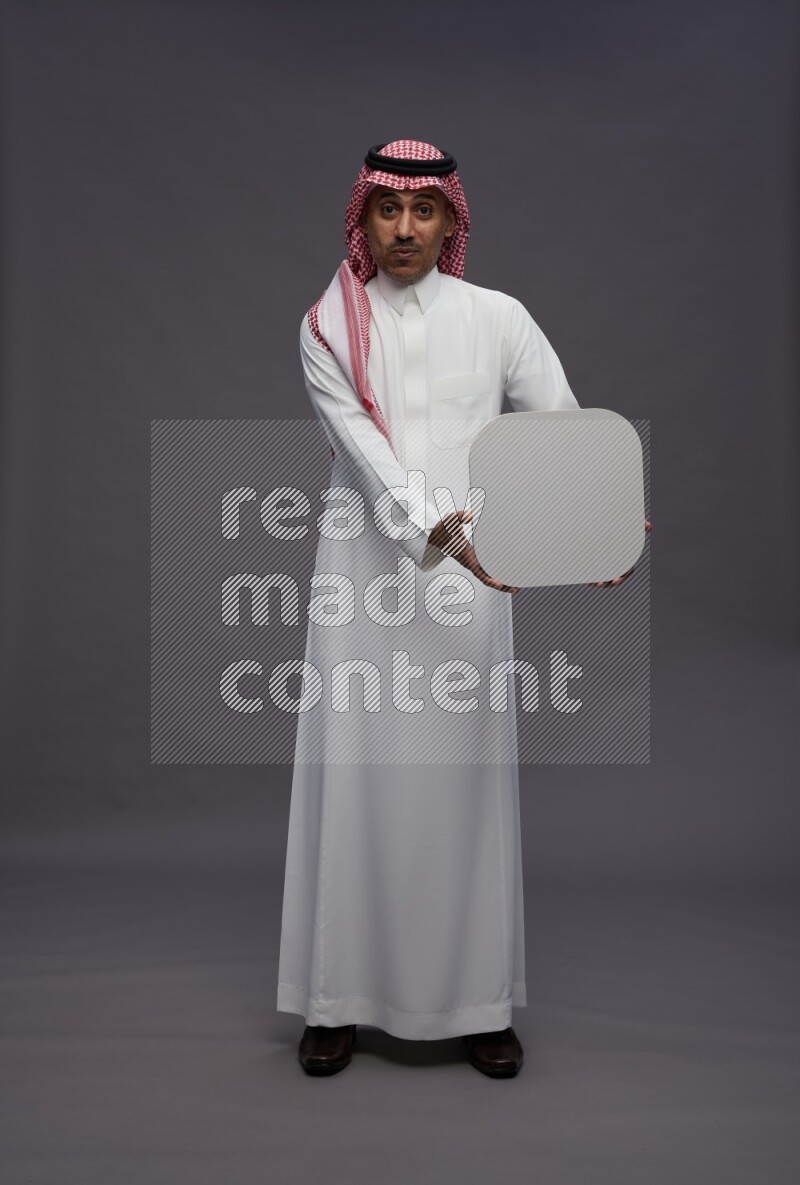 Saudi man wearing thob and shomag standing holding social media sign on gray background