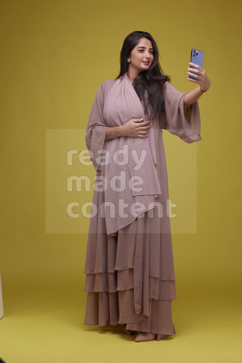 A woman Taking a Selfie on a Yellow Background wearing Brown Abaya