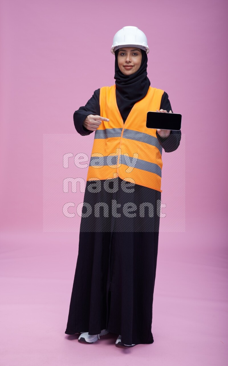 Saudi woman wearing Abaya with engineer vest and helmet standing showing phone to camera on pink background