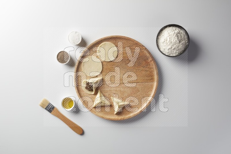 two closed sambosas and one open sambosa filled with meat while flour, salt, black pepper and oil with oil brush aside in a wooden dish on a white background