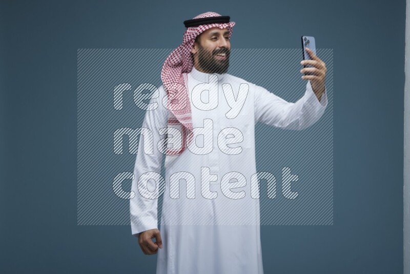 A man Taking Selfie in a blue background wearing Saudi Thob with Shomag
