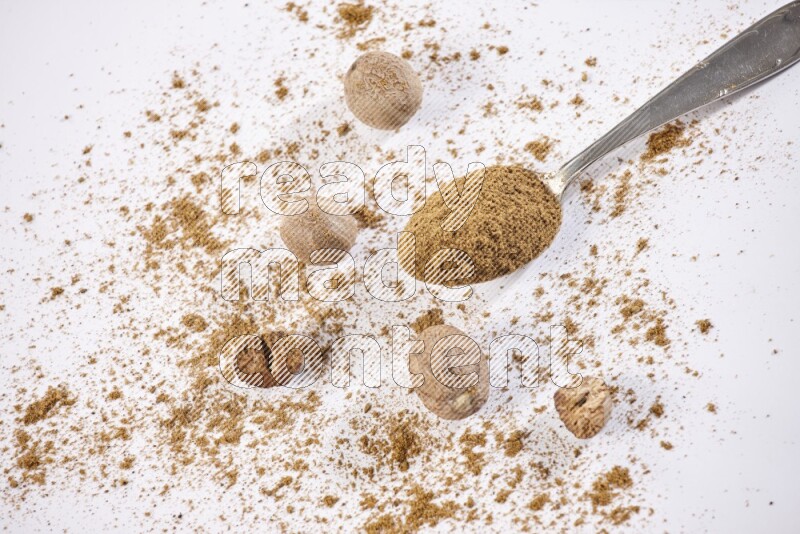 A metal spoon full of nutmeg powder with some of the powder and seeds beside it on a white flooring