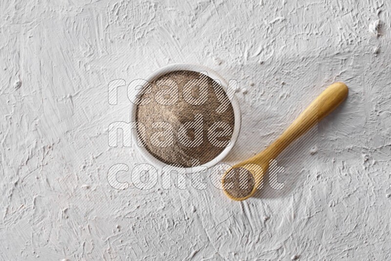 A pottery white bowl full of black pepper powder and wooden spoon full of powder on textured white flooring