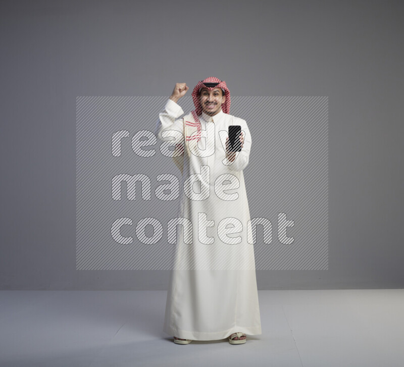 A Saudi man standing wearing thob and red shomag showing phone to camera on gray background
