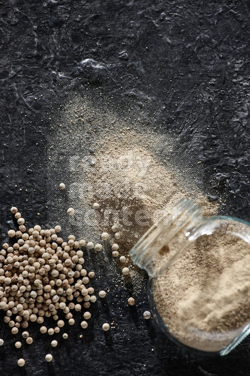 A flipped herbal glass jar full of white pepper powder with spilled powder and pepper beads on textured black flooring