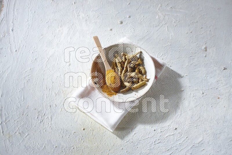 A plate filled with dried turmeric whole fingers and a wooden spoon full of turmeric powder on a textured white flooring