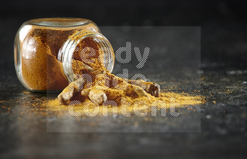 A flipped glass spice jar full of turmeric powder and powder spilled out of it with dried whole fingers on textured black flooring