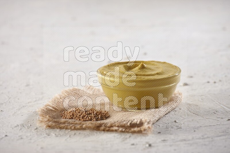 A glass bowl full of mustard paste set on a burlap piece with some mustard seeds spread on a textured white flooring