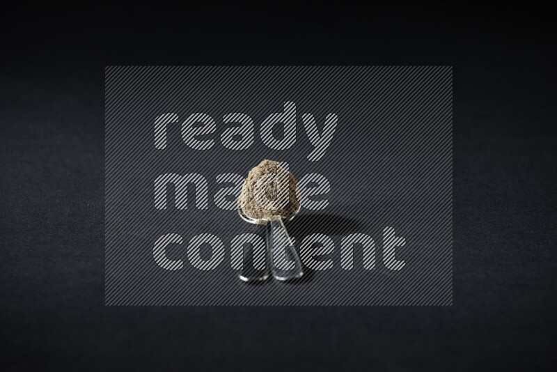 2 Metal spoons full of cardamom powder on black flooring