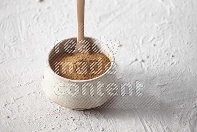 Ceramic beige bowl full of cinnamon powder with a wooden spoon on a textured white background