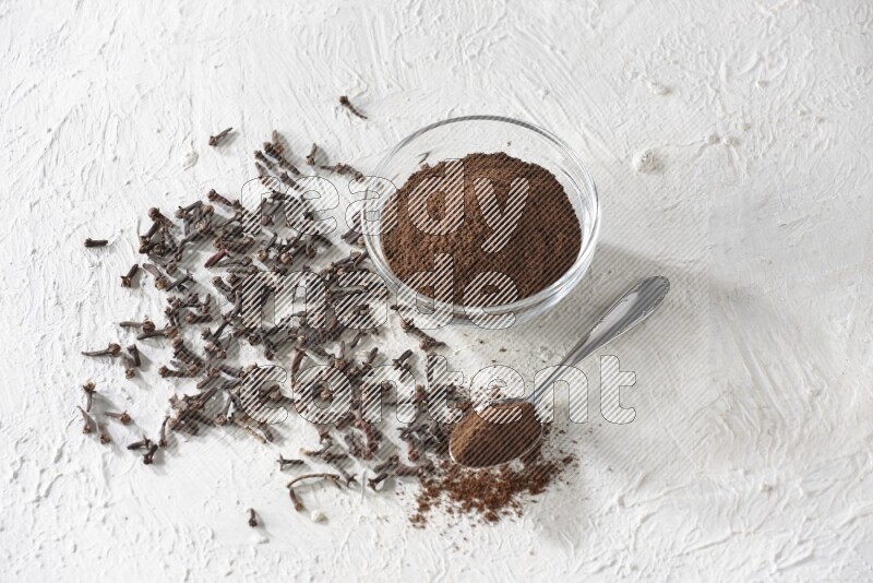 A glass bowl and a metal spoon full of cloves powder with cloves grains spread on a textured white flooring