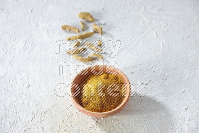 A wooden bowl full of turmeric powder with dried whole fingers on textured white flooring