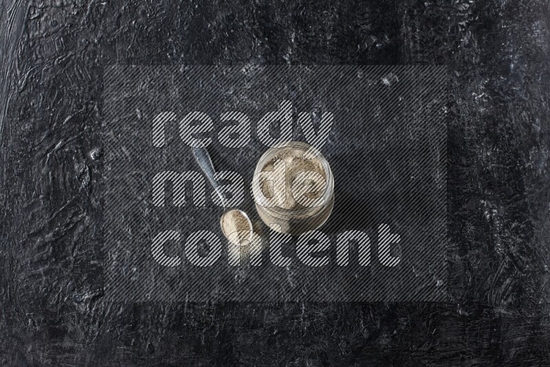 A glass jar and a metal spoon full of white pepper powder on textured black flooring