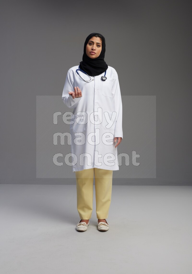 Saudi woman wearing lab coat with stethoscope standing interacting with the camera on Gray background