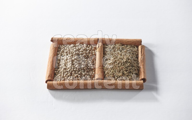 مربعان من أعواد القرفة ممتلئان بحبوب الفلفل الأبيض وحبوب الكمون علي خلفية بيضاء