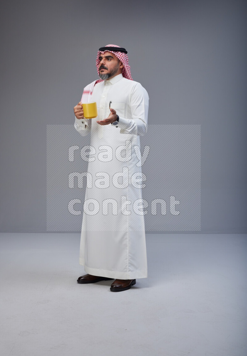 Saudi man Wearing Thob and red Shomag standing holding mug on Gray background